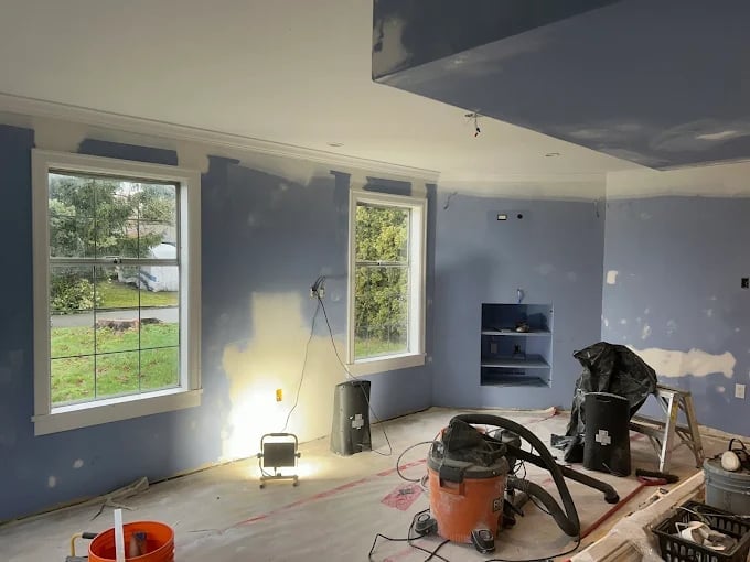 Interior of a home under construction with blue drywall, large windows, and various tools and equipment scattered throughout