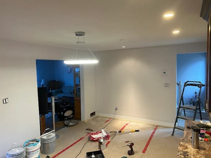 Interior construction site with exposed floor, red masking tape layout, hanging light fixture, and partially finished room with blue doorway